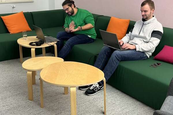 Zwei Männer sitzen mit zwei Laptops auf einem grünen Sofa. 