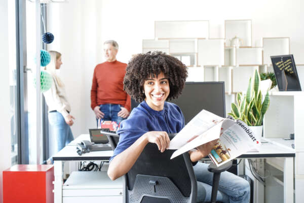 Frau mit Zeitschrift in der Hand sitzt an Bildschirmarbeitsplatz.
