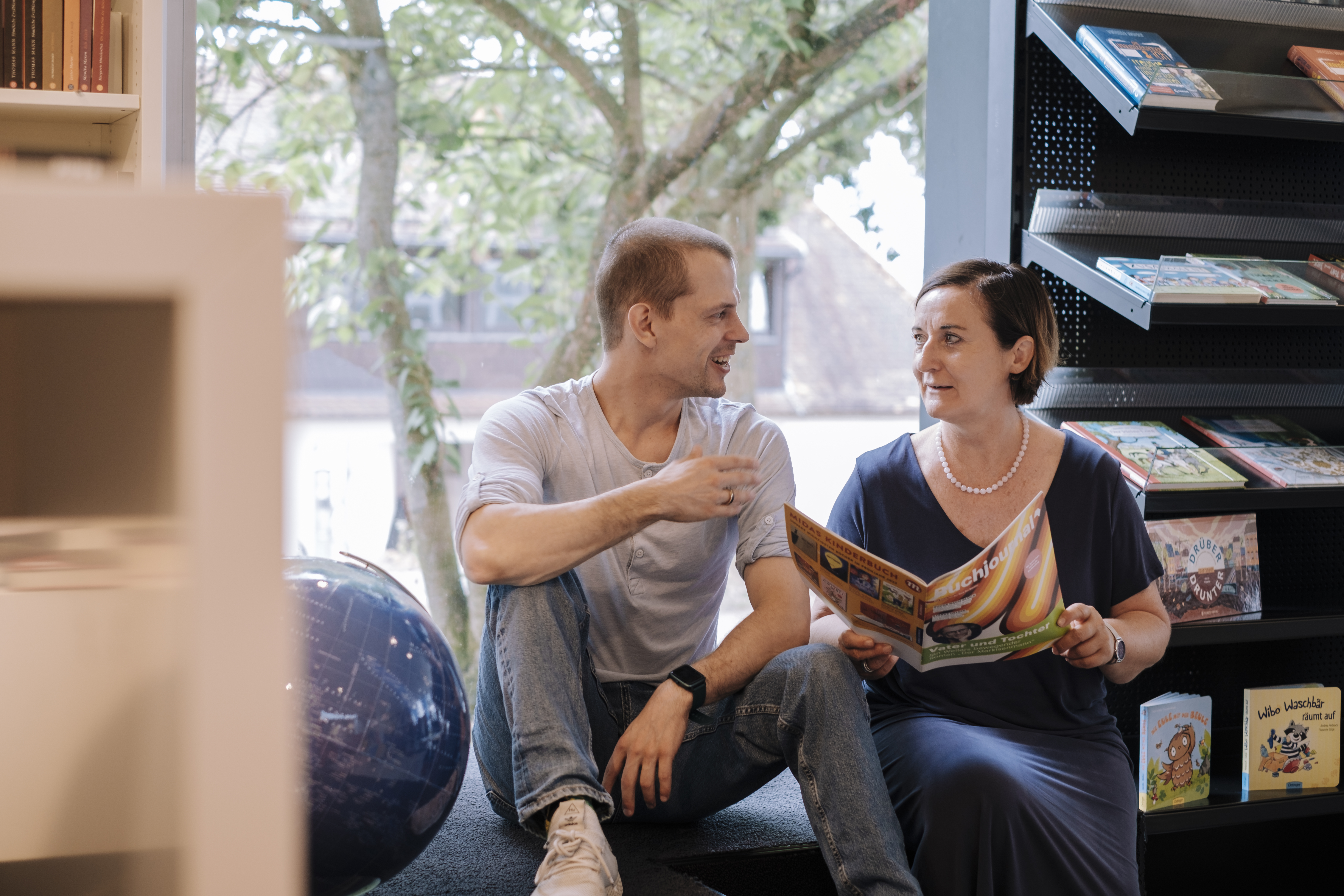 Ein Mann und eine Frau sitzen in einer Buchhandlung und sprechen miteinander. Die Frau hält ein Buchjournal in den Händen.