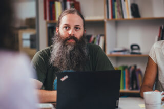 Ein Mann, der besonders im Kamerafokus liegt, sitzt mit weiteren Personen an einem Tisch mit seinem Laptop und unterhält sich.