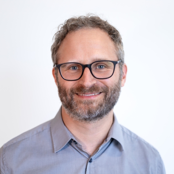 Johannes steht vor einer weißen Wand und trägt ein hellblaues Hemd.