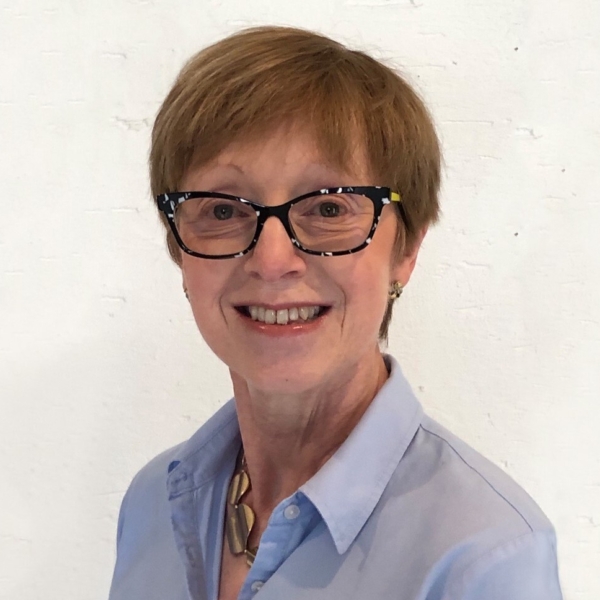 Portrait von Liz McNaughton die in die Kamera lächelt. Sie trägt eine Brille mit schwarzem Gestell und eine blaue Bluse.