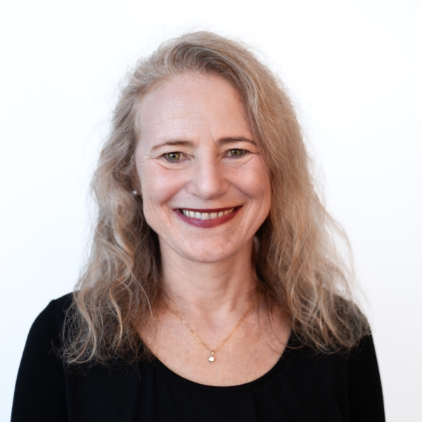 Portrait von Susanne Fuchs, die in die Kamera lächelt und vor einer weißen Wand steht.