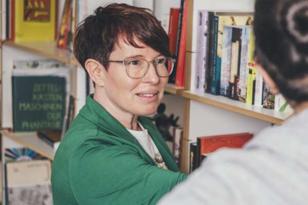 Silke erklärt einer Person etwas vor eine Bücherregal