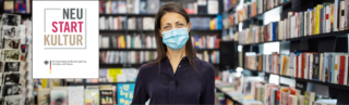 Eine Frau steht trägt einen Mundschutz und steht vor einem Bücherregal. Neben ihr ist ein Plakat mit der Aufschrift "Neu Start Kultur".