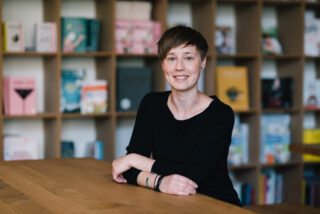 Kathrin Riechers sitzt an einem Tisch vor einem Bücherregal und lächelt.