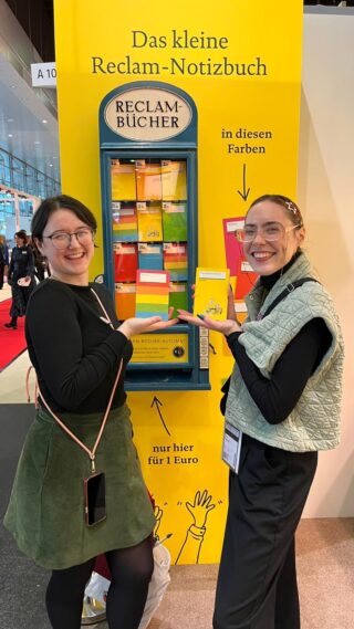 Franziska Werum und Lorina Bischoff stehen vor einem Reclam-Automaten und halten Reclam-Notizbücher