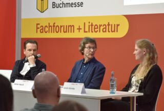 Stefan Hauck, Carolin Reintke und ein weiterer Mann tragen Mikrofone und sitzen an einem Tisch. Im Hintergrund steht Fachforum + Literatur.