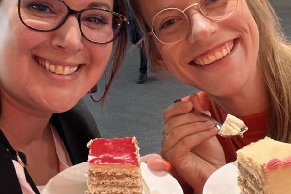 Katrin und Angie halten jeweils einen Teller mit einem Stück der Jubiläumstorte in den Händen und lächeln.