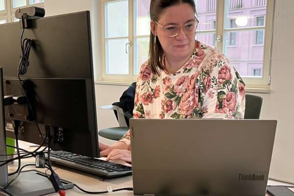 Eine Frau sitzt an einem Tisch vor einem Laptop und einem Computerbildschirm.