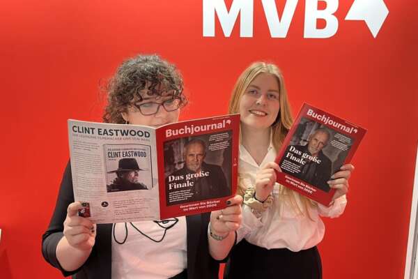 Vanessa und Olivia stehen nebeneinander vor einer roten Wand und schauen jeweils in ein Buchjournal Magazin.