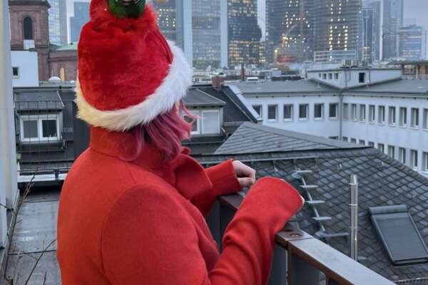 A person stands on a railing and looks out over the Frankfurt skyline. She is wearing a red coat and a red Christmas hat. 