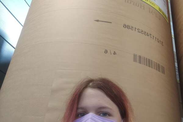 Selfie of Anna standing with a mask on her face in front of a huge roll of paper in a print shop. 