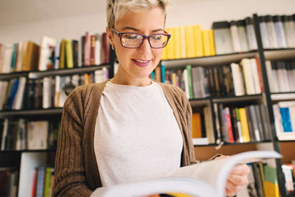 Eine Frau steht vor einem Bücherregal und sieht sich lächelnd ein Buch an.