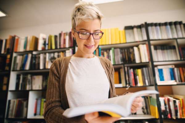 Eine Frau steht vor einem Bücherregal und sieht sich lächelnd ein Buch an.