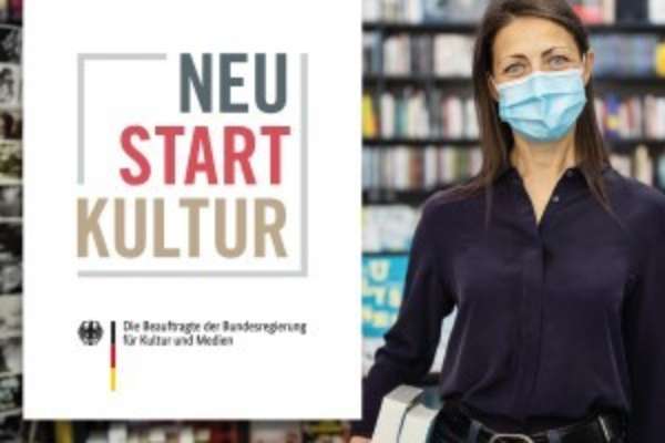 Eine Frau steht trägt einen Mundschutz und steht vor einem Bücherregal. Neben ihr ist ein Plakat mit der Aufschrift "Neu Start Kultur".