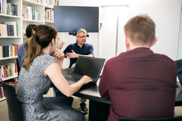 Eine Gruppe von Menschen sitzt mit Laptops an einem Tisch und unterhält sich.