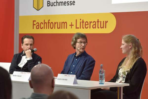 Stefan Hauck, Carolin Reintke und ein weiterer Mann tragen Mikrofone und sitzen an einem Tisch. Im Hintergrund steht Fachforum + Literatur.