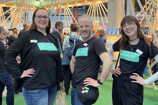 Drei Personen stehen mit passenden Gastlands-T-Shirts vor dem Gastland Messestand.