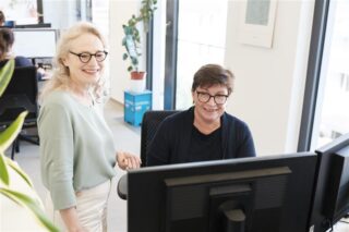 Eva Singer sitz vor einem Computerbildschirm, Susanne Fuchs steht daneben. Beide schauen in die Kamera.